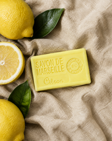 Savon de Marseille citron jaune sur un lin gris sauge, avec des citrons entiers, un demi-citron et des feuilles vertes