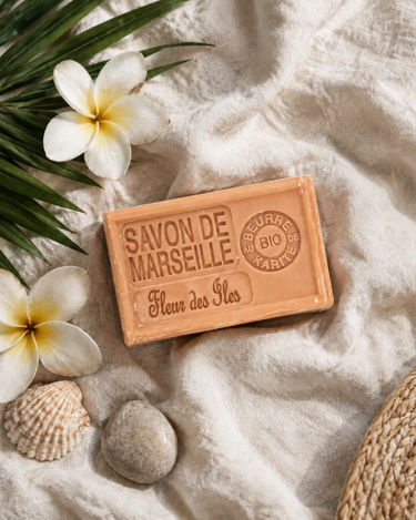 Savon de Marseille Fleur des Îles sur un lin blanc cassé, avec des fleurs de frangipanier, une feuille de palmier et un coquillage