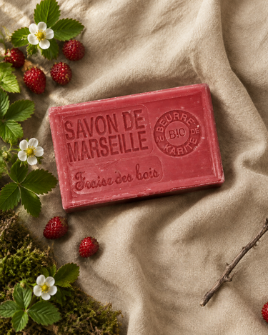 Savon de Marseille fraise des bois sur un lin beige, avec de petites fraises sauvages, des fleurs blanches, de la mousse et une brindille