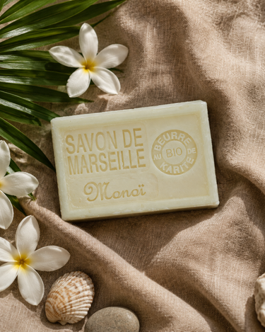 Savon de Marseille monoï sur un lin beige, avec des fleurs de tiaré blanches, une feuille de palmier et un coquillage